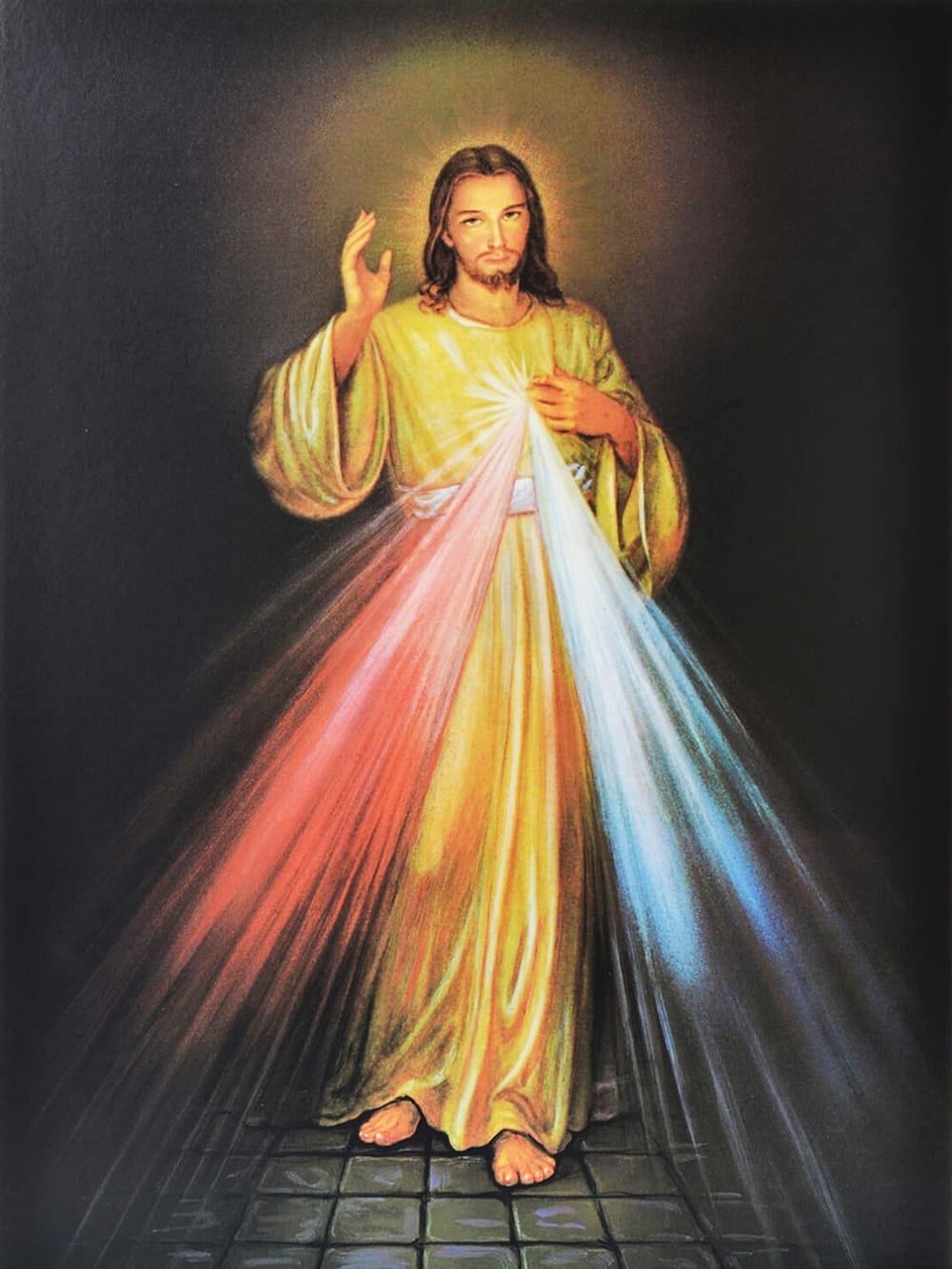 A devotional painting of Jesus Christ known as the Divine Mercy. Christ stands with his right hand raised in blessing and his left hand pointing to his heart. Two rays of light, one red and one pale, stream downward from His heart, symbolizing blood and water. He is dressed in a flowing white robe, and His expression is serene and compassionate, inviting trust and devotion. The background is simple, emphasizing the radiant light emanating from His heart.